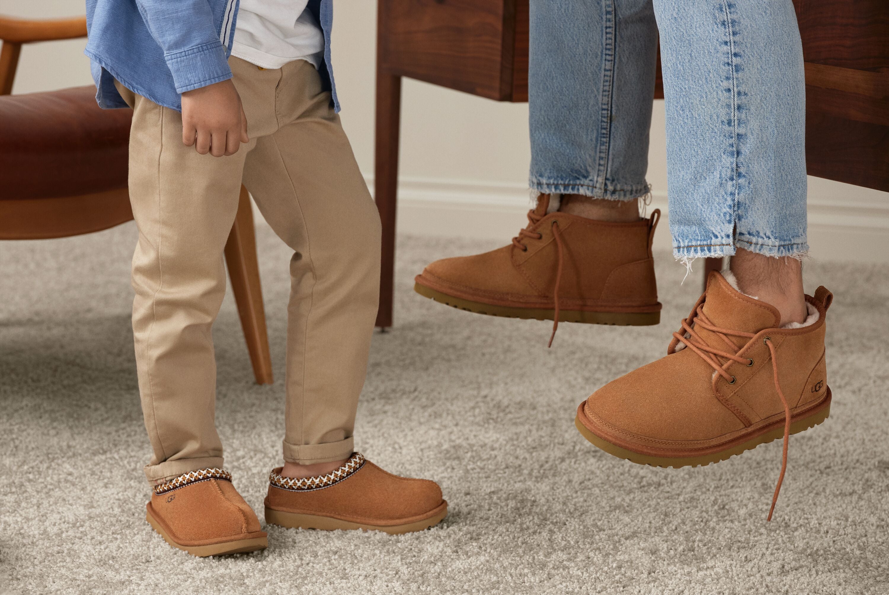 A man and a boy stand on a rug in front of a chair, both wearing brown shoes and sharing a moment together.