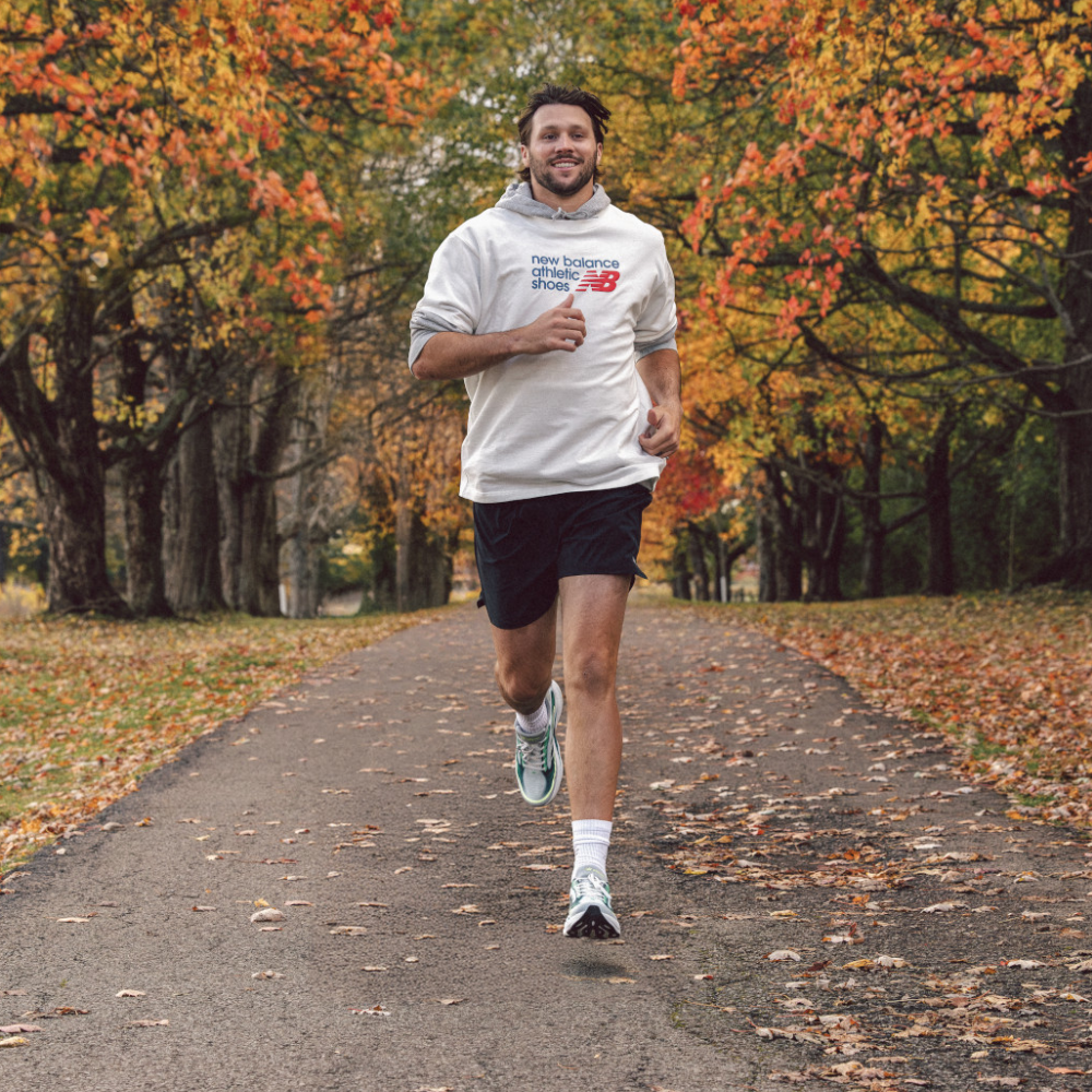 Josh Allan running on a leaf-covered path in an autumn park, wearing a white New Balance hoodie, black running shorts, and the New Balance Ellipse