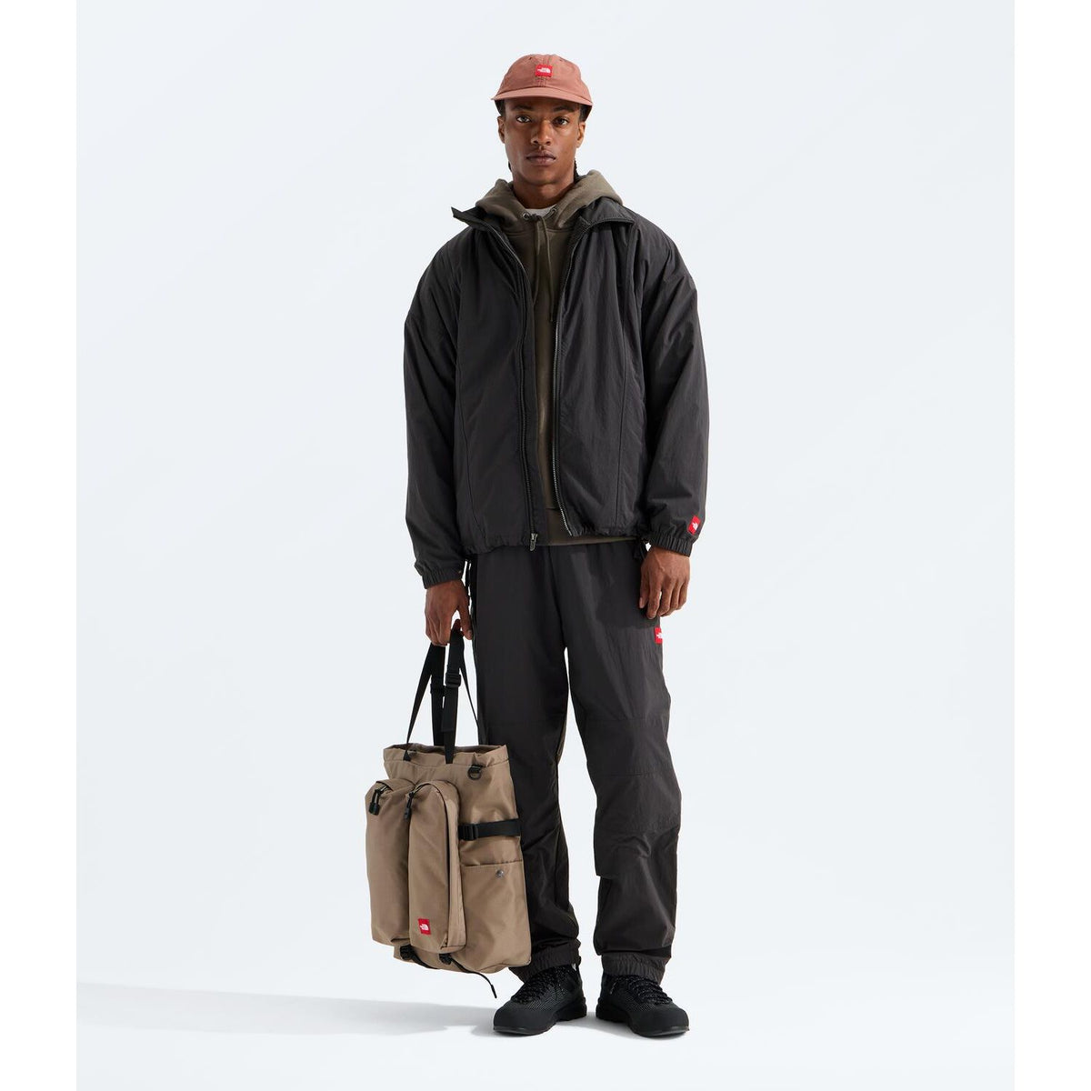 Man wearing a black nylon WindWall jacket with a Red Box logo patch, matching trousers, and a tan tote bag.