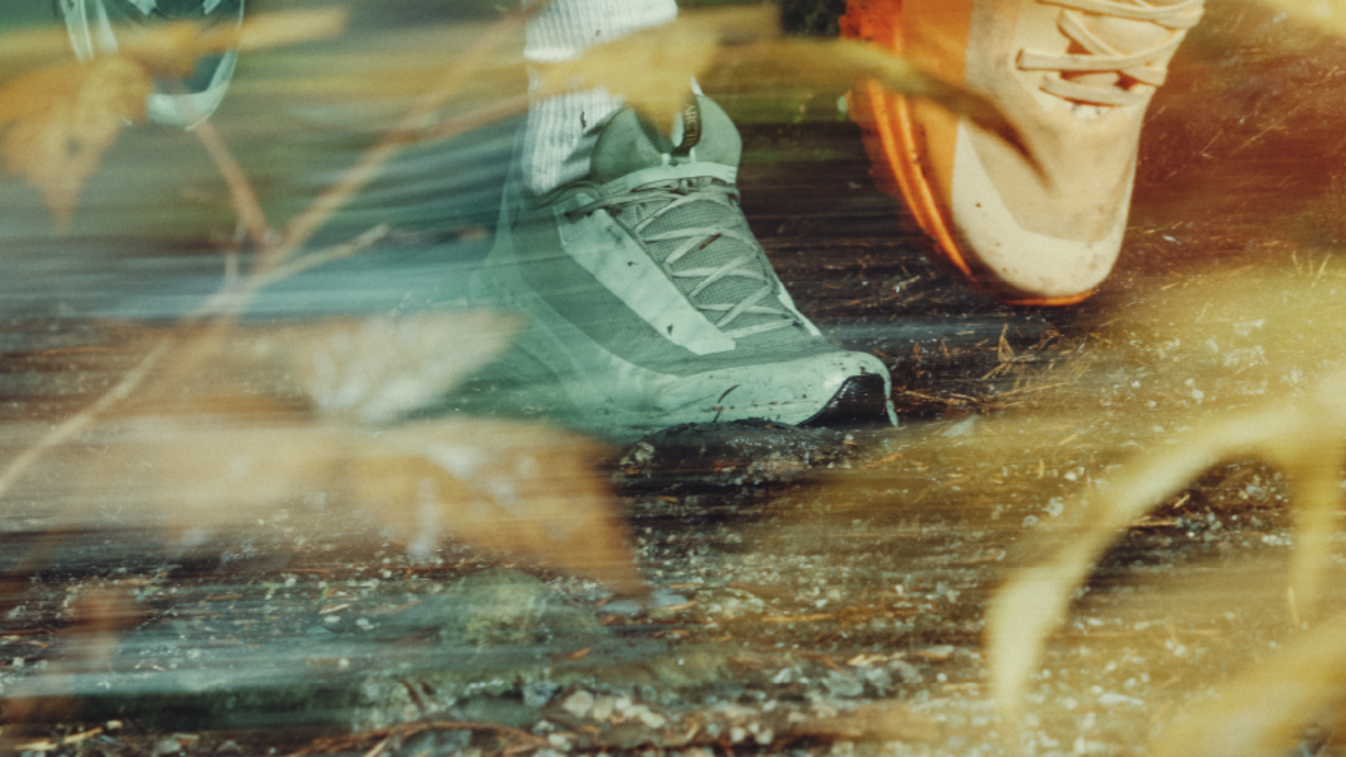 Close-up of two people running on a trail, with one wearing grey and the other wearing orange shoes. Motion blur and autumn leaves are in the foreground.