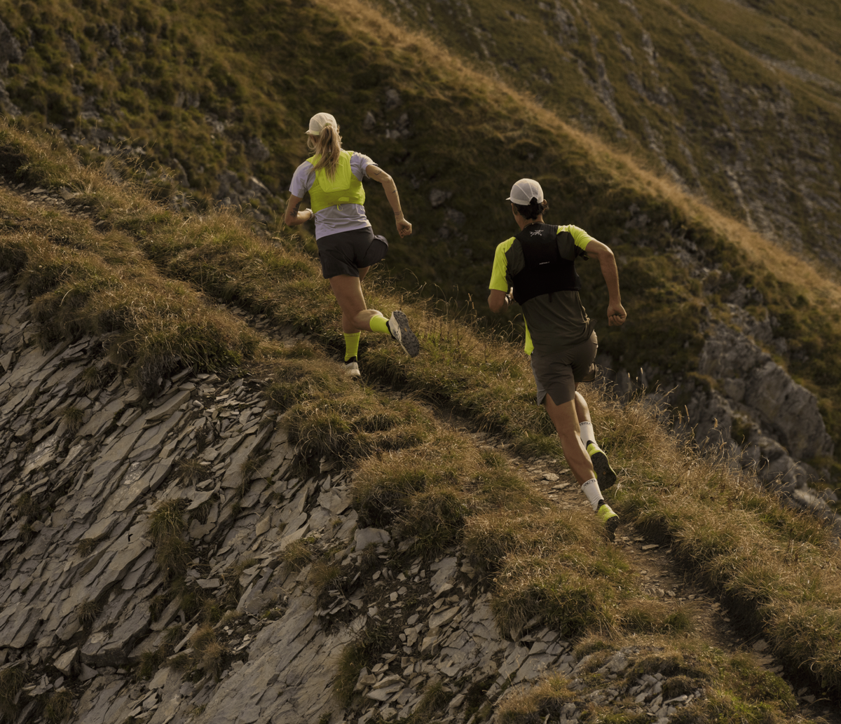 Two trail runners climbing a steep grassy mountain slope, shown from behind. Both runners wearing Arc'teryx Trail Running Gear including the new Sylan 2